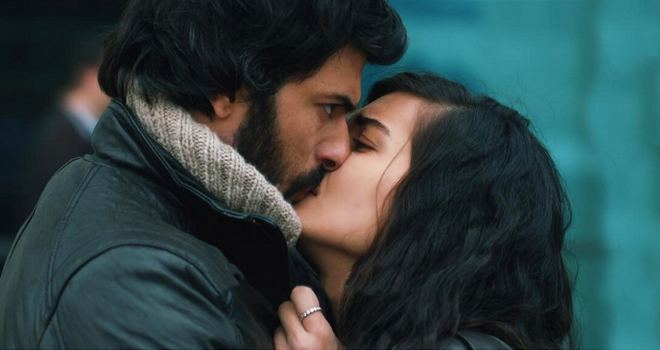 Engin Akyürek kissing a woman while wearing a black jacket and gray scarf