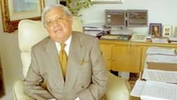 Enrique Molina Sobrino sitting on a chair while wearing a brown coat and a tie inside a room