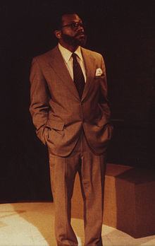 Charles S. Dutton as Dysart in Equus, looking afar and wearing eyeglasses, brown coat, white long sleeves, necktie, and brown pants