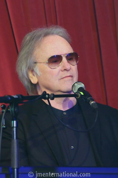 Eric Stewart with a microphone in front of him while he is wearing a black coat, long sleeves, and brown sunglasses