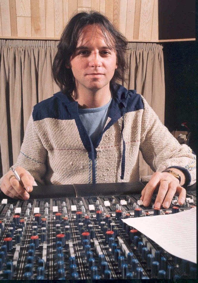 Eric Stewart with a tight-lipped smile while holding the audio mixer and wearing a light blue shirt underneath a cream and blue jacket