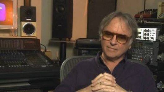 Eric Stewart sitting on the chair inside the studio with his hands together while he is wearing blue long sleeves and sunglasses