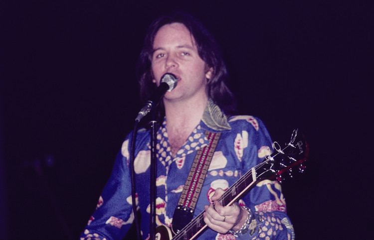 Eric Stewart singing while playing the guitar and wearing a blue and white long sleeves