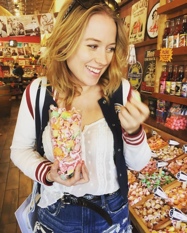 Erica Deutschman smiling while holding a bottle of candies, with short blonde hair, with sunglasses on her head, wearing a white, black and red jacket over a white shirt and blue jeans.