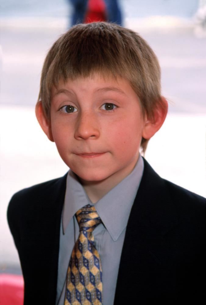 Eric Per Sulivan is smiling, standing, has brown hair wearing a light blue polo with gold and blue printed necktie under the black coat.