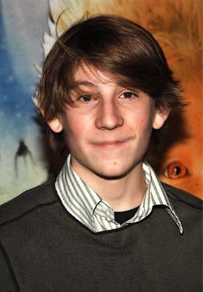 Eric Per Sulivan is smiling, standing in front of the printed backdrop, has brown hair, black eyes, wearing a black shirt under a striped white polo and black long sleeve on top.