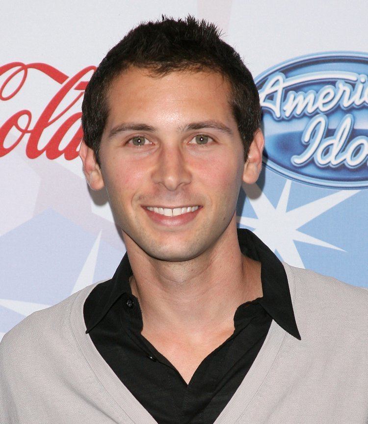 Eric Per Sulivan is smiling, standing in front of american idol backdrop, mouth half open, has brown hair, brown eyes, shaved mustache and beard wearing a black polo under a gray long sleeve shirt.