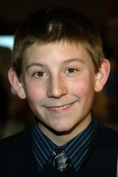 Eric Per Sulivan is smiling, has brown hair, brown eyes, wearing a dark blue striped polo and necktie under the black coat.