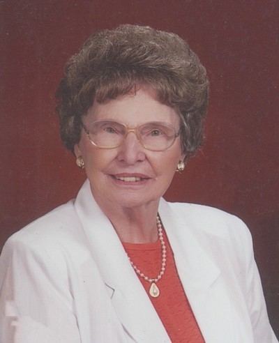 Ernestine Wiedenbach smiling, with curly short hair, wearing eyeglasses, earrings, necklace, and a white blazer with an orange shirt.