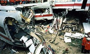 Eschede derailment, 1998 in Germany. A white broken train with a red line on the side. Broken pieces of parts on the ground. Two people wearing red orange suits with black pants and white helmets.