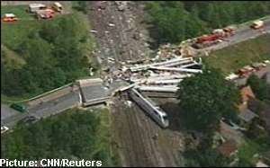 A helicopter shot of the Eschede derailment in 1998 in Germany, The white train with red line color on the side derailed and crashed. Broken bridge. Cars and fire trucks on the top left side. Houses on the right side. Green Trees around the road.