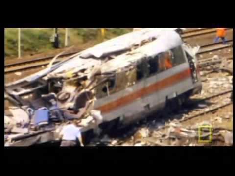 Eschede derailment, 1998 in Germany. A part of white broken train with a red orange line on the side. Broken pieces of parts on the ground. In the middle of the railroad. on the left a man checking the broken train wearing a white polo and black pants.