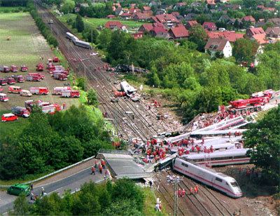 Eschede derailment in 1998 in Germany, The white train with red line color on the side derailed and crashed. Broken bridge with a group of people wearing red suits. Cars and fire trucks on the left side. Houses on the right side. Green Trees around the road.