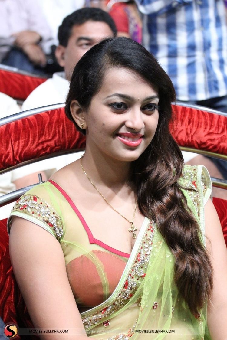 Ester Noronha smiling and sitting on a red chair with black wavy hair and bindi on her forehead while wearing earrings, a cross necklace, a red and yellow-green beaded see-through saree