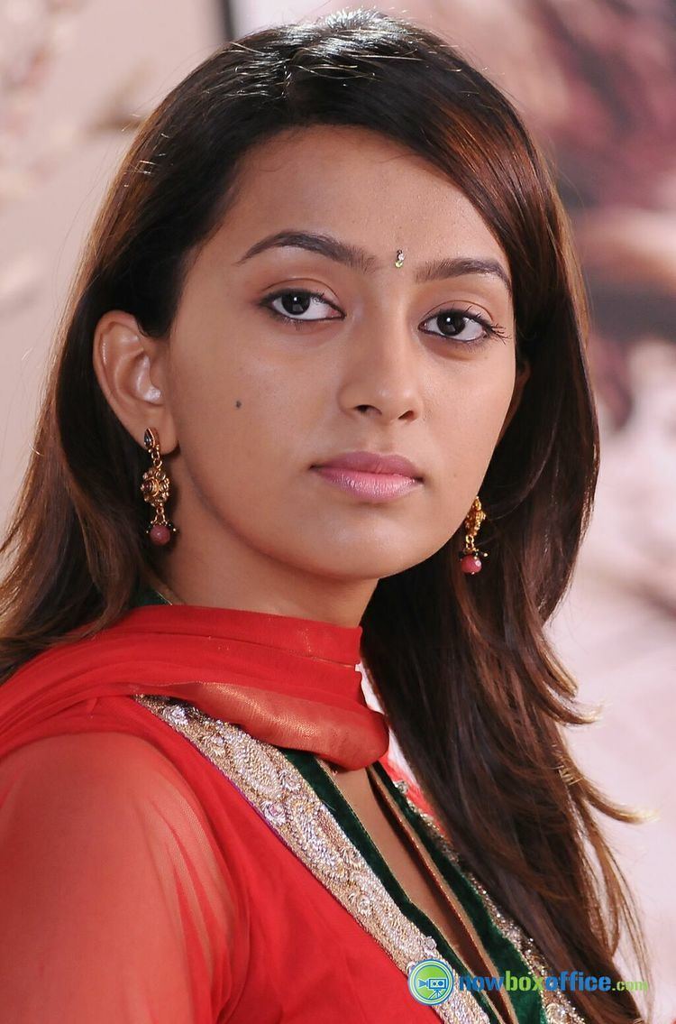 Ester Noronha with a serious face, wavy hair, and a diamond bindi on her forehead while wearing earrings, a red scarf, and a red dress with a black and light beige neckline