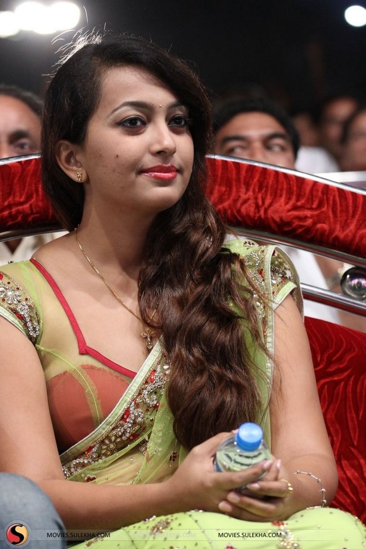 Ester Noronha smiling, sitting on a red chair, and holding bottled water, with wavy hair and bindi on her forehead while wearing earrings, a cross necklace, bracelet, ring, and a red and yellow-green beaded see-through saree