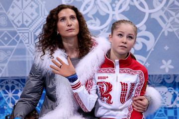 Eteri Tutberidze and Yulia Lipnitskaya are looking above with serious faces. Eteri with curly hair and wearing a gray coat with a fur design while Yulia is waving her hand and wearing a white and red jacket.