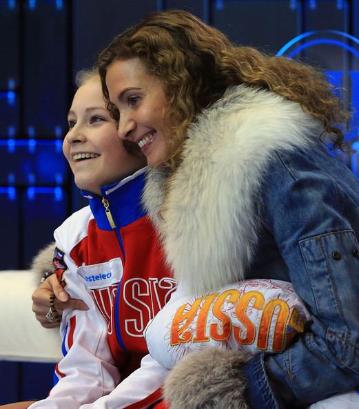 Eteri Tutberidze and Yulia Lipnitskaya are smiling. Eteri with curly hair and wearing a denim jacket with a fur design while Yulia is wearing a multi-colored jacket.