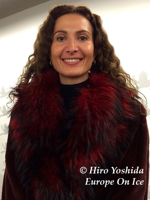 Eteri Tutberidze smiling, with curly hair, wearing earrings, and a maroon fur coat.