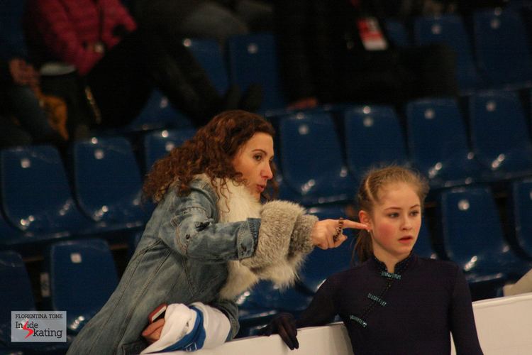 Eteri Tutberidze pointing at something with Yulia Lipnitskaya with a serious face inside a skating rink. Eteri with curly hair and wearing a denim jacket with a fur design while Yulia is wearing a black unitard.