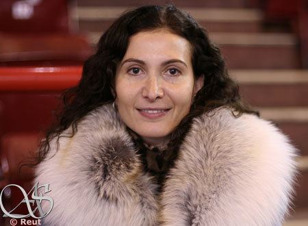 Eteri Tutberidze smiling, with curly hair, wearing earrings, and a fur coat.