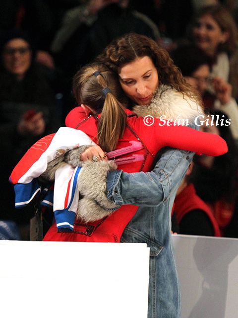 Eteri Tutberidze and Yulia Lipnitskaya hugging each other. Eteri with a sad face, curly hair, and wearing a denim jacket with a fur design while Yulia is wearing a red unitard.