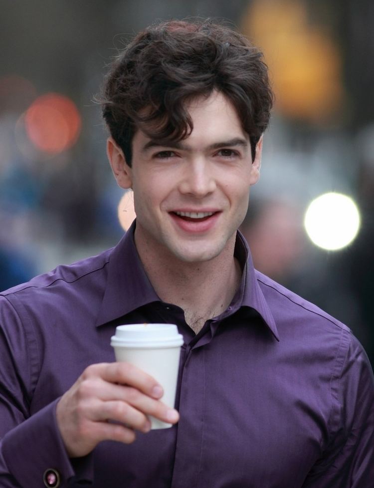 Ethan Peck is smiling while holding a white cup of coffee in his right hand, he has black hair, chest hair, and wearing a purple polo long sleeve