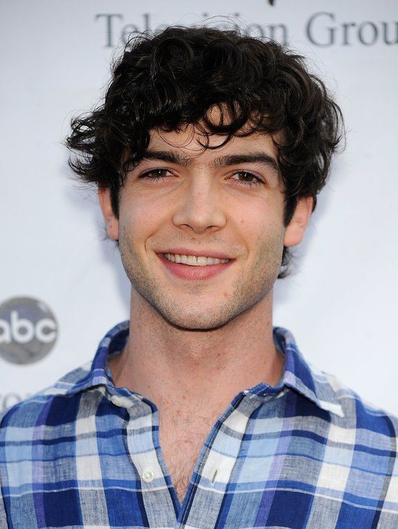 Ethan Peck is smiling with his black hair, chest hair, beard, and mustache, and wearing a blue checkered polo