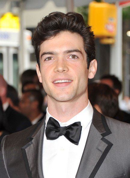 Ethan Peck is smiling and has black hair with people in the background, wearing a white polo with a black bow tie under a black coat