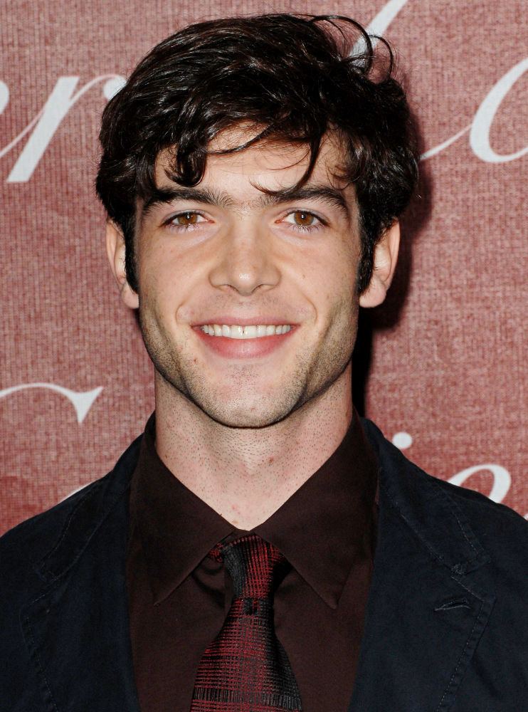 Ethan Peck is smiling and has black hair, beard and mustache, wearing a black polo long sleeve with a maroon neck tie