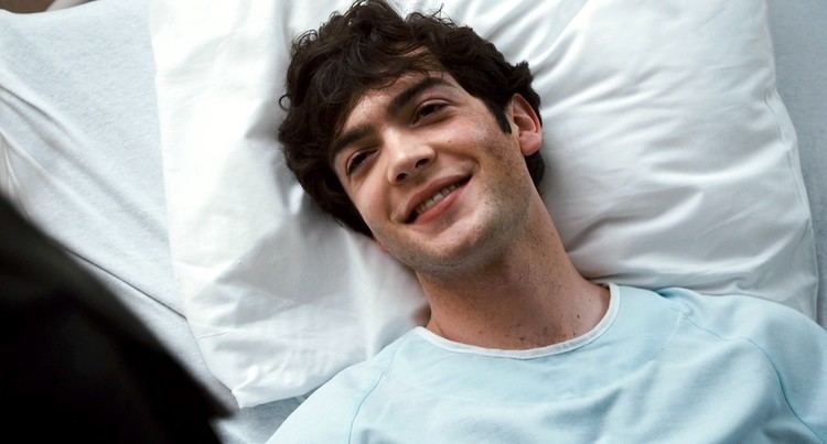 Ethan Peck is smiling while lying on a white pillow on a white bedsheet, with his black hair, and wearing a blue shirt
