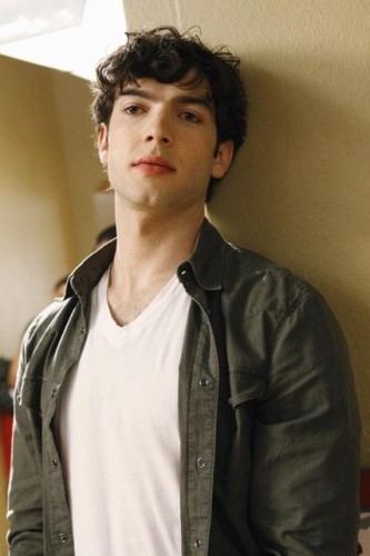 Ethan Peck seriously looking at something while leaning his head and back on a wall, he has black hair and wearing a white shirt under a black polo long sleeve