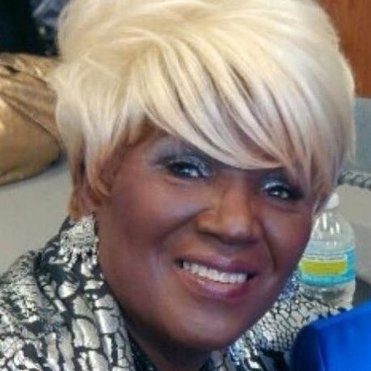 Etterlene DeBarge smiling with a blonde wig and wearing black and gray clothes.