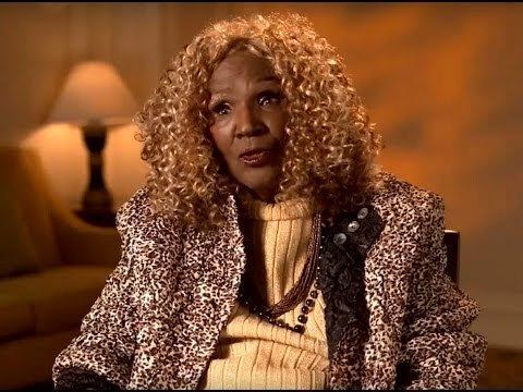 Etterlene DeBarge talking in an interview with longer hair and wearing a spotted coat over yellow clothes.