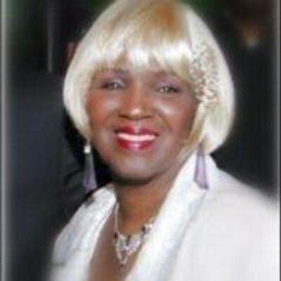 Etterlene DeBarge smiling and wearing a blonde wig along with a white dress and some jewelry.