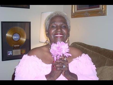 Etterlene DeBarge smiling while holding a pink flower and wearing a white dress off shoulder dress.