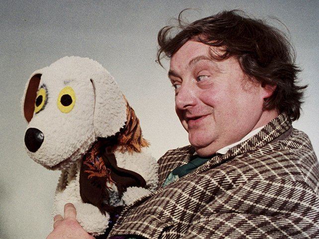 Eugene Lambert playing with a dog puppet, wearing a white shirt under a brown coat and a green necktie