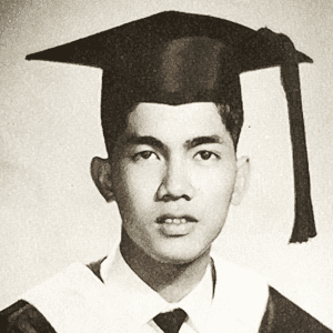 Evelio B. Javier smiling, wearing a black graduation hat, a black and white graduation robe over white long sleeves, and a black tie.