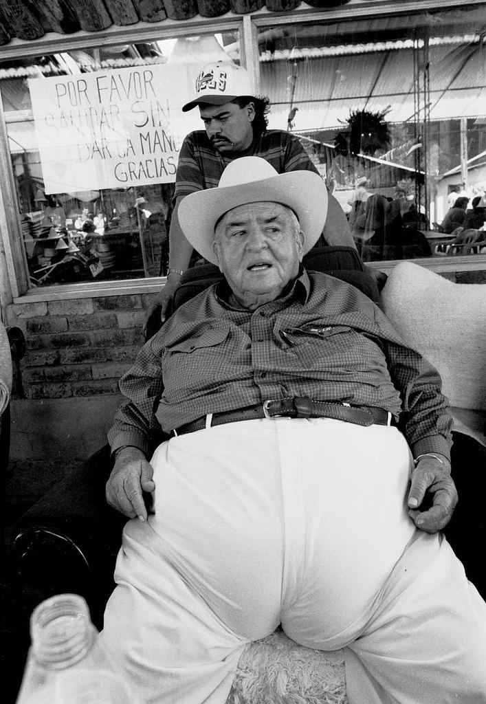 Fabio Ochoa Restrepo wearing a hat, checkered long sleeves, and white pants while sitting on a couch with a man at his back wearing a cap and a striped polo shirt.