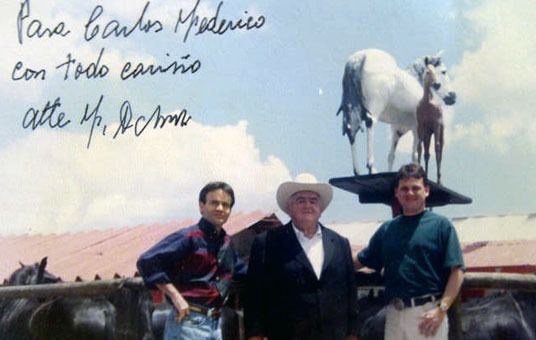 Fabio Ochoa Restrepo in the center wearing a black coat, white long sleeves, white hat, and the two men beside him wearing blue and red long sleeves and a green shirt with horses at their back.