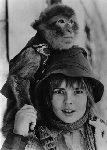 Young Fabrice Josso smiling and carrying a monkey while wearing a hat, scarf, and jacket