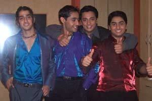 On the left, Qazi Touqeer is smiling, standing with both hands in his pocket, having black wavy hair, wearing a necklace, blue shirt, dark blue coat, black belt, and black pants. 2nd from left, a man, smiling, standing while looking to his left with his hand in his pocket, he has black hair and is wearing a blue long sleeve, a black belt, and black pants. 3rd from left, Rex D'Souza is smiling, standing with his right arm on top of a man (left), right-hand thumbs up, left arm on the shoulder of Arijit Singh (right), left-hand thumbs up, has black hair, and is wearing a black long sleeve, and on the right, Arijit Singh, smiling, standing with both arms up and thumbs up, has long black hair, wearing a red long sleeve and black pants.