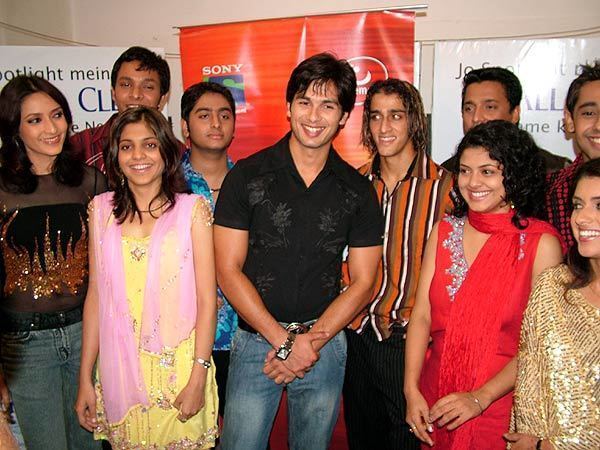 From the contestants of Fame Gurukul 2005, from left: Arpita Mukherjee is smiling, standing looking to her left, she has long black hair and is wearing a black see-through shirt with a flame print on it and denim pants. 2nd from left at the back is a man smiling, standing with black hair, wearing a red shirt with black stripes. In front of him, 3rd from left, is Monica Gadgil, smiling, standing with her hands on her sides, wearing a yellow dress and a pink see-through scarf. 4th from left, at the back, Arijit Singh is serious, has black hair, and is wearing a blue flower-printed polo with a black necklace and black pants. 5th from left, is Shahid Kapoor, smiling, standing, while his right hand holds the left arm, he has black hair and is wearing a black floral printed polo, a silver watch, a black belt, and denim pants. 6th from left, Qazi Touqeer is smiling, standing looking at Shahid, has long wavy hair with brown highlights, and is wearing striped pants and an orange polo with maroon, black, and white stripes. 7th from left, at the back, is a man standing with black hair and wearing a black shirt. In front of him, 8th from left is Mona Bhatt, smiling, with black wavy hair, wearing earrings, a red scarf, a gold bracelet, rings, and a red dress with a white silver design. On the right side, at the back, is a man smiling, standing, with brown hair, wearing a dotted red polo, and in front of him is a woman smiling, with brown hair, wearing a gold shiny long sleeve dress.
