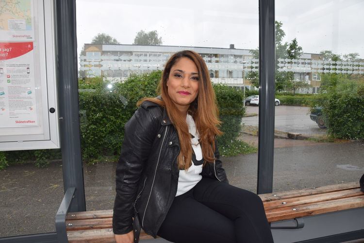 Farah Abadi is smiling while sitting on a bench, with long blonde hair, wearing a black leather jacket over a white shirt and black pants.