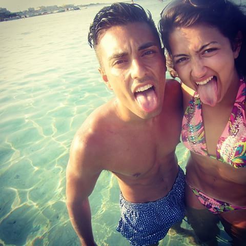 Farah Abadi and Oscar Zia are showing their tongues at the beach. Farah is wearing a multi-colored swimsuit white Oscar is topless and wearing blue shorts.