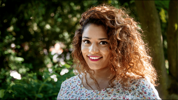 Farah Abadi smiling, with curly blonde hair, with trees in the background. Farah is wearing a multi-colored top.
