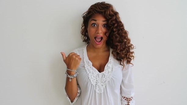 Farah Abadi is smiling with an open mouth and pointing his thumb to the right, with curly hair and visible cleavage, wearing bracelets and a white long sleeve top.