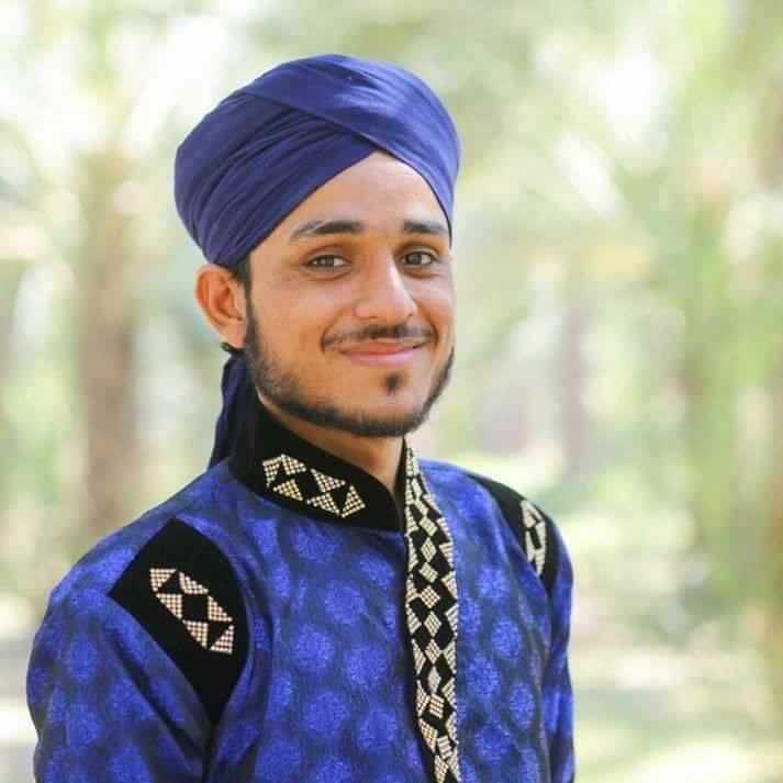 Farhan Ali Qadri smiling and wearing a blue turban and clothing with black and white patterns with small facial hair.