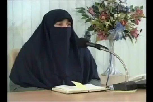 Farhat Hashmi with sad-looking eyes with an open book on the top of the table and a microphone, wearing a gray thobe and a black Shemagh.