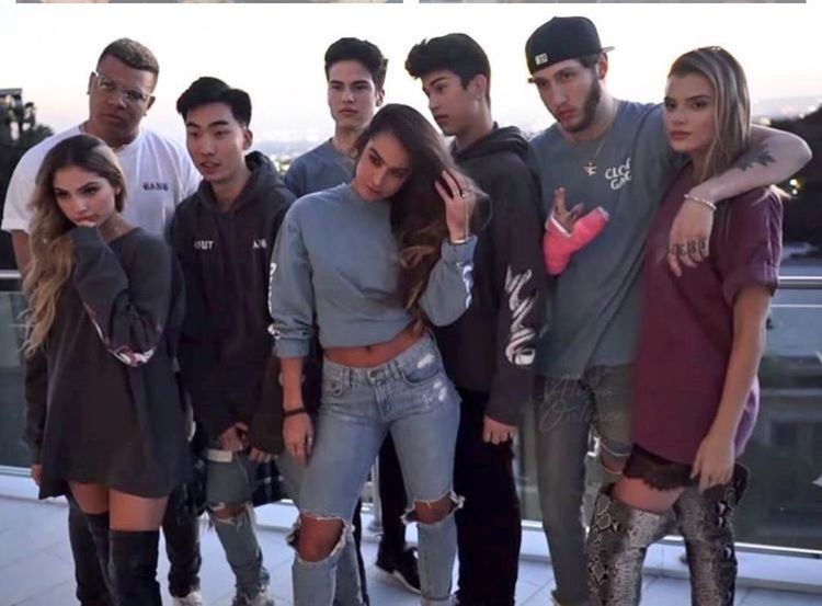 FaZe Banks looking serious with arms around the neck of a girl and wearing a gray shirt, jeans, and a cap during a photoshoot with the Clout Gang members